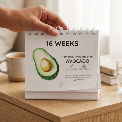 A hand holds a spiral-bound 16-week baby milestone desk calendar showing an avocado graphic and text comparing the baby's size to an avocado. The calendar sits on a wooden table with a coffee cup, books, and a glass of water in a softly lit room.